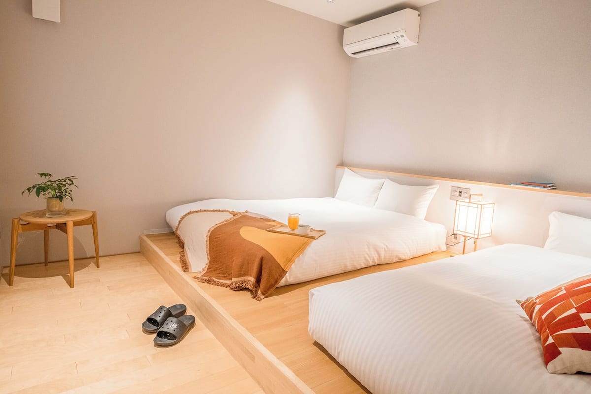 A hotel room with two beds, a small table with a plant, a pair of slippers, and a bedside lamp.