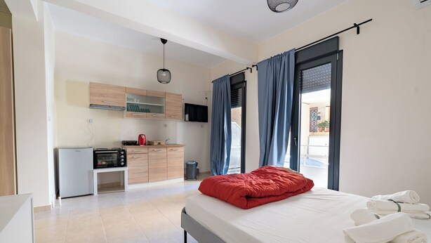 A modern bedroom with a bed, white linens, and a red pillow. A small kitchenette is visible in the background with a microwave, toaster, and cabinets. - Ag. Ioannis m. station 1 bed for 2 pers by MPS (Vironas)
