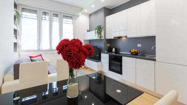 A modern kitchen with white cabinets, a black countertop, and a dining area with a glass table and white chairs. - notaMi - Monte Ceneri Suite (Milan)