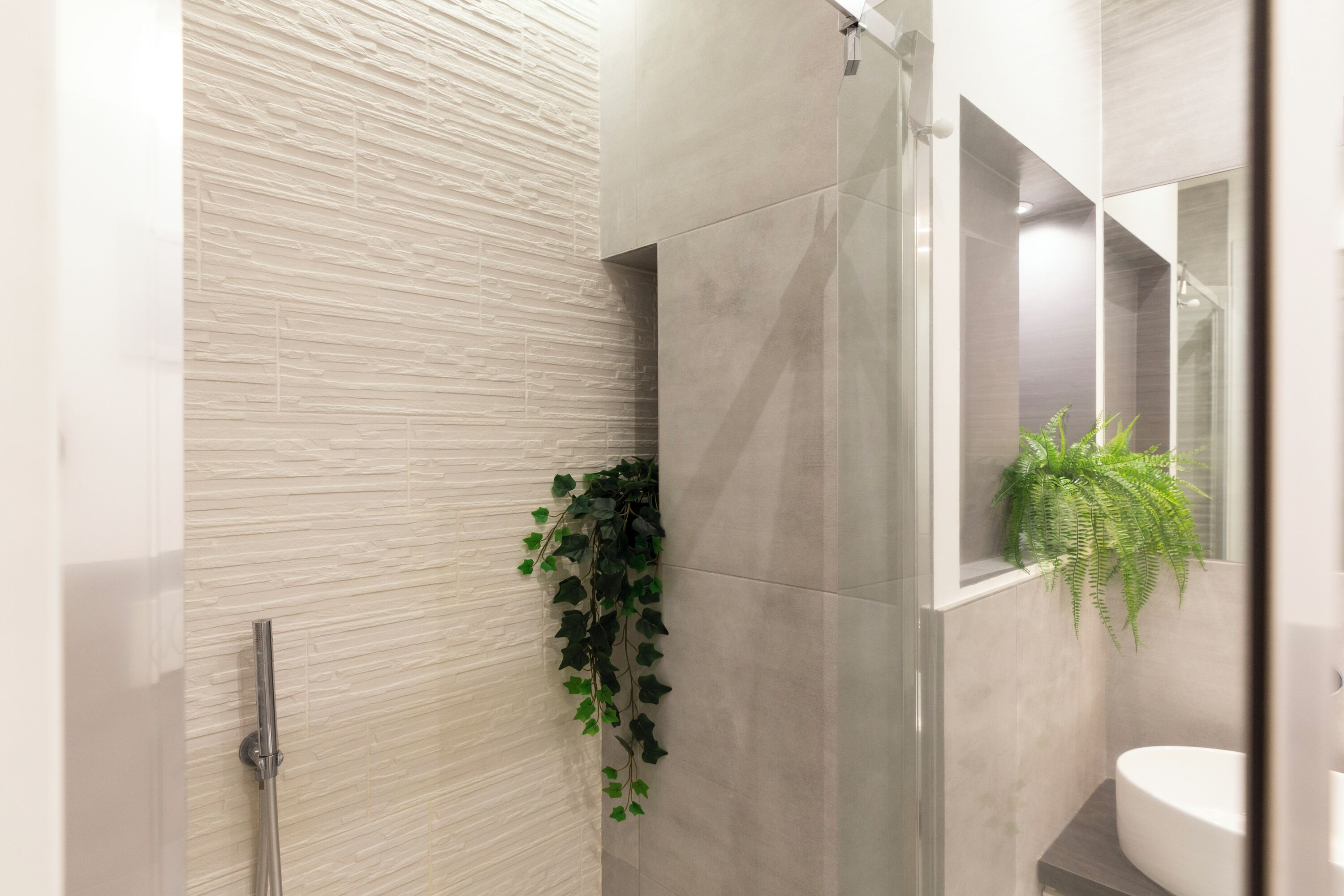 A modern bathroom with a textured wall, a glass shower enclosure, a sink with a mirror, and a potted plant.