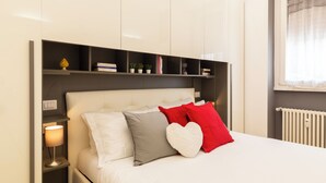 A neatly arranged bedroom with a bed, bedside tables, and a shelf with books and plants. - notaMi - Monte Ceneri Suite (Milan)