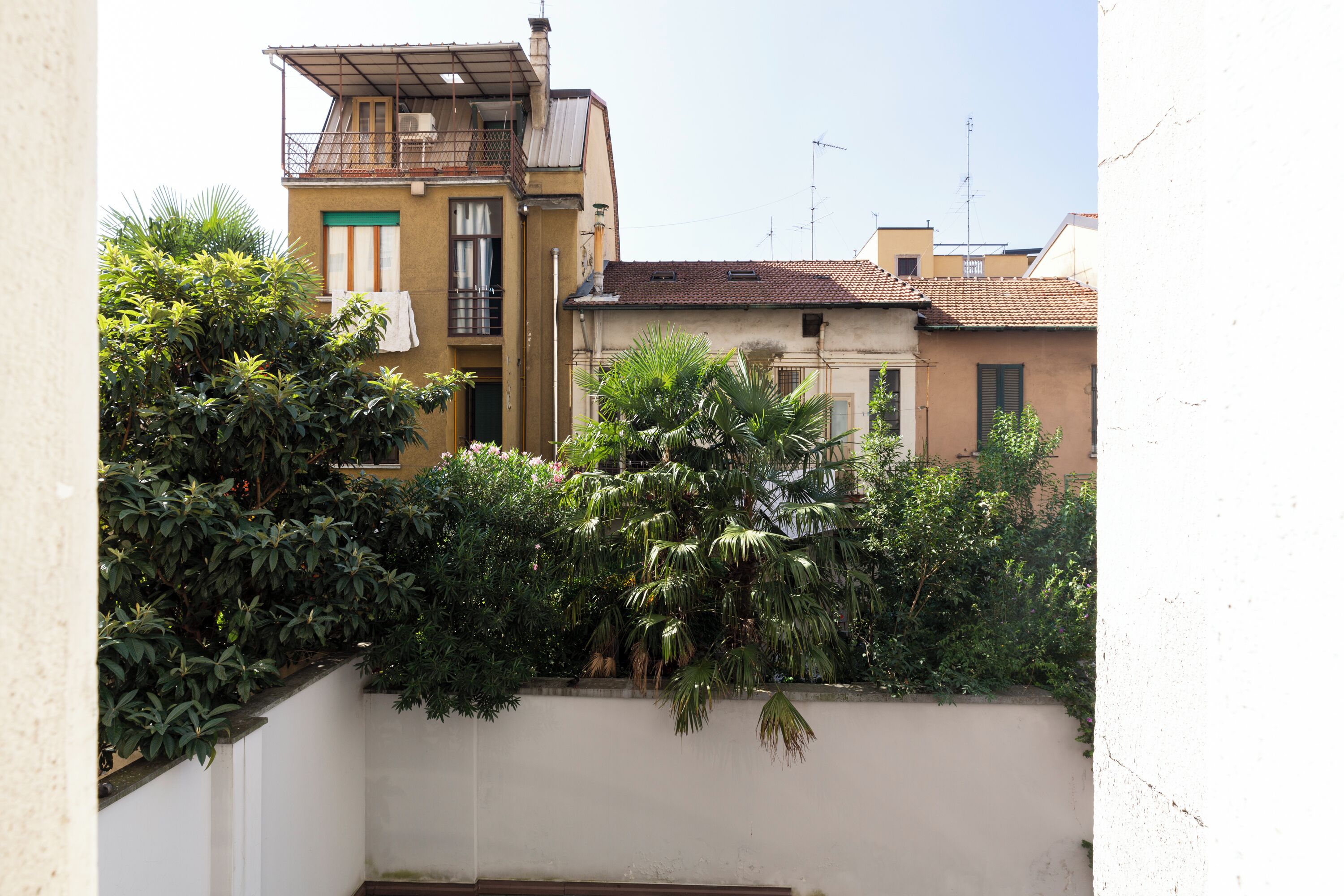 A residential building with a balcony, a white wall, and dense greenery in front.