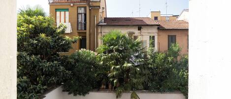 A residential building with a balcony, a white wall, and dense greenery in front.
