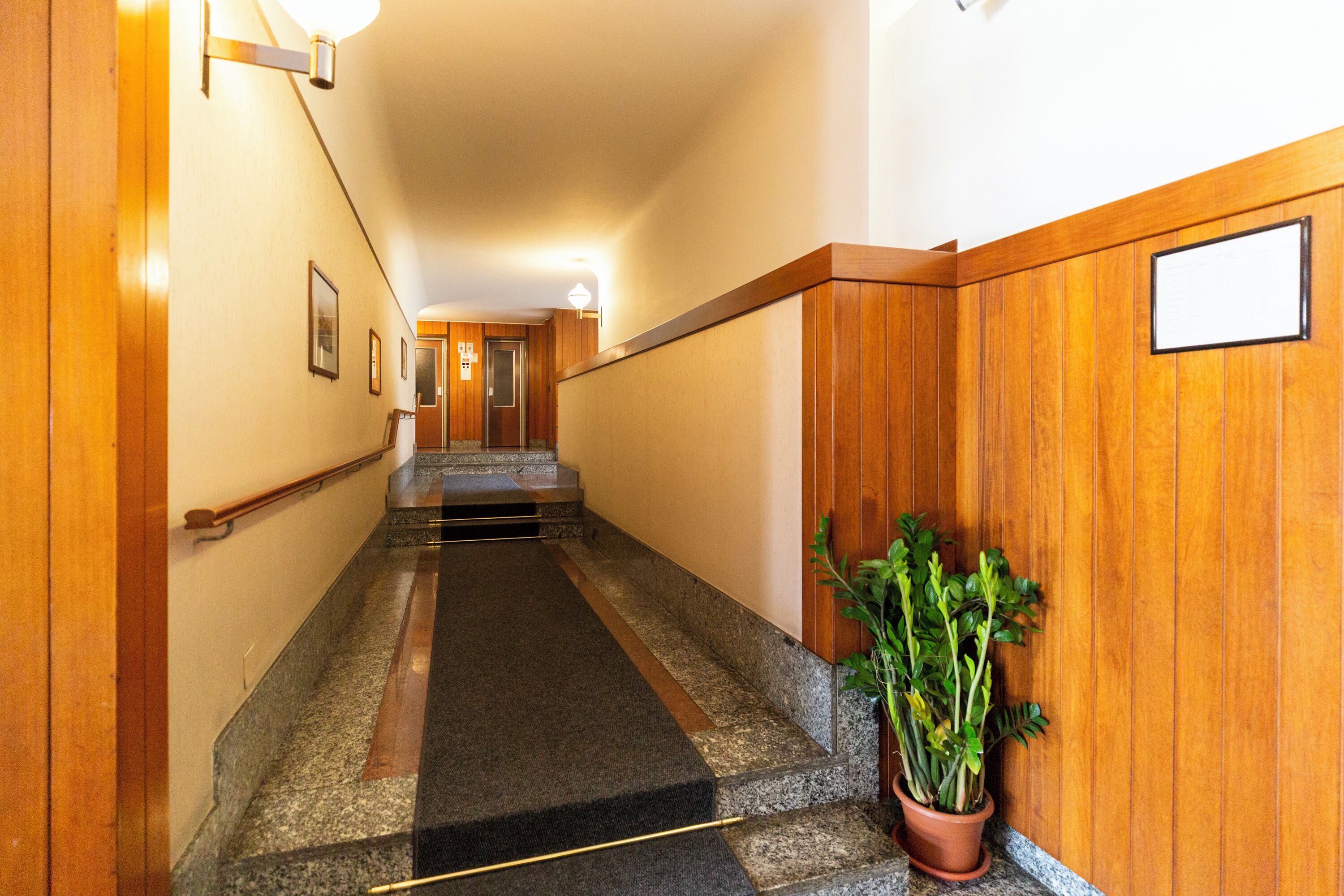 Um corredor com painéis de madeira, uma planta em vaso e uma escada que leva a um elevador.