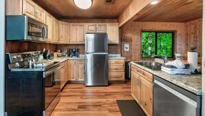 A modern kitchen with wooden cabinets, stainless steel appliances, granite countertops, and a large window offering a view of greenery.