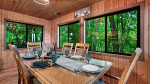 A wooden cabin dining area with a glass table, chairs, and large windows offering a forest view.