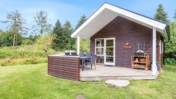 A small wooden cabin with a covered patio, a dining area with chairs, and a fireplace.