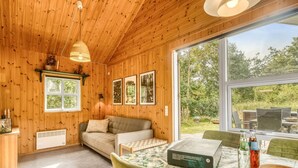 A cozy cabin interior with wooden walls, a sofa, a dining table with chairs, and a large window offering a view of the outdoors.