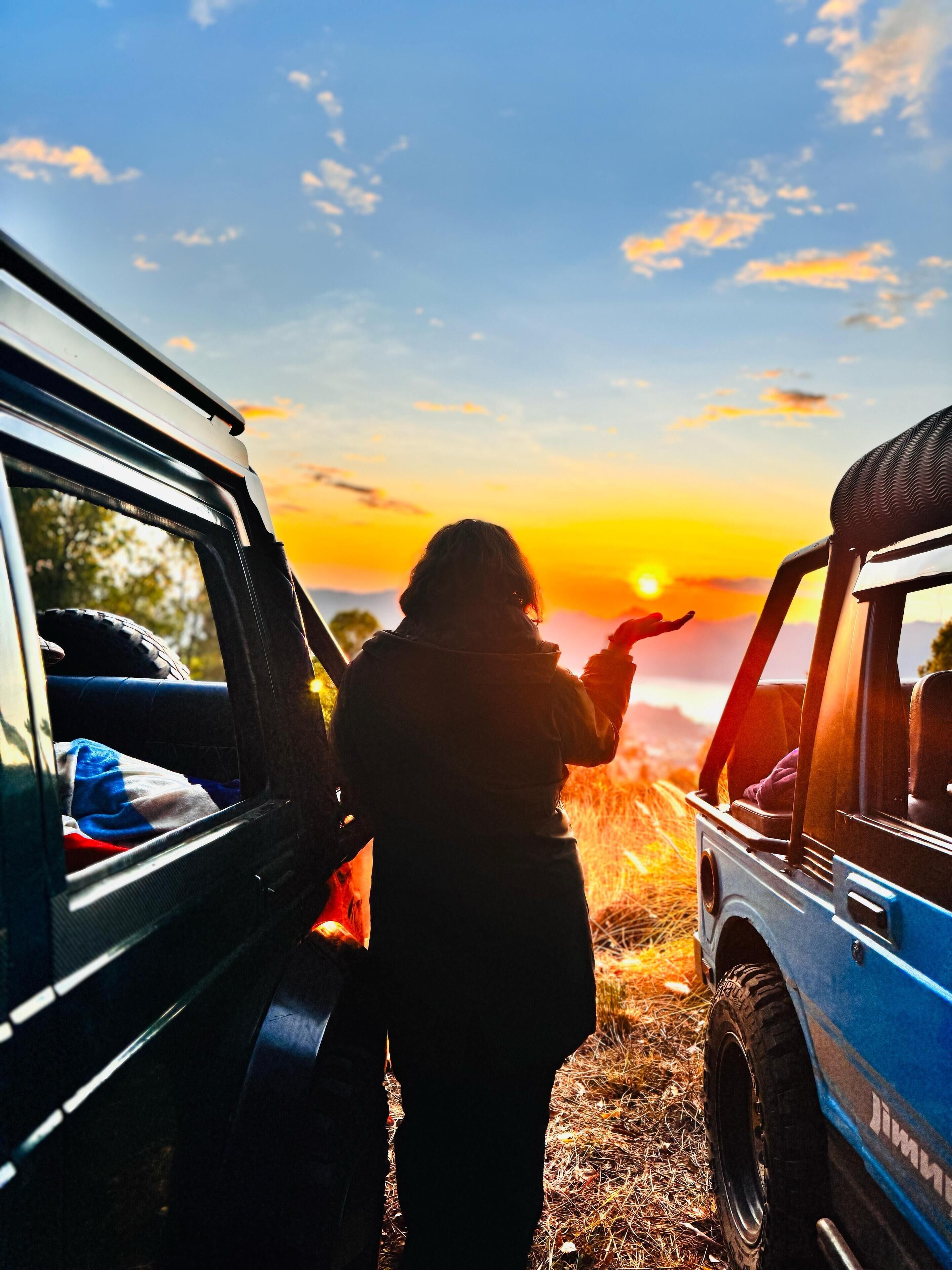 A person standing between two vehicles, one blue and one black, with a sunset in the background.