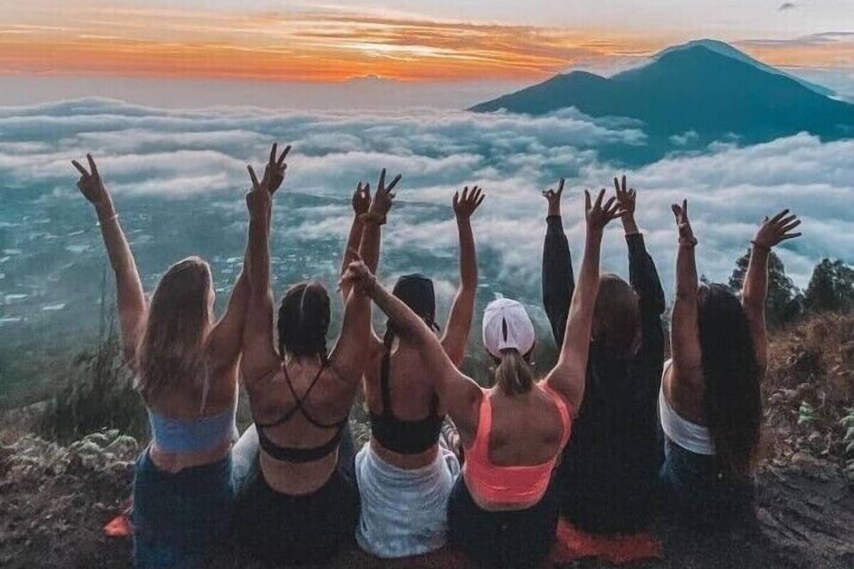 Un grupo de personas sentadas en una montaña, de espaldas a la cámara, contemplando una puesta de sol sobre un paisaje nublado con otra montaña al fondo.