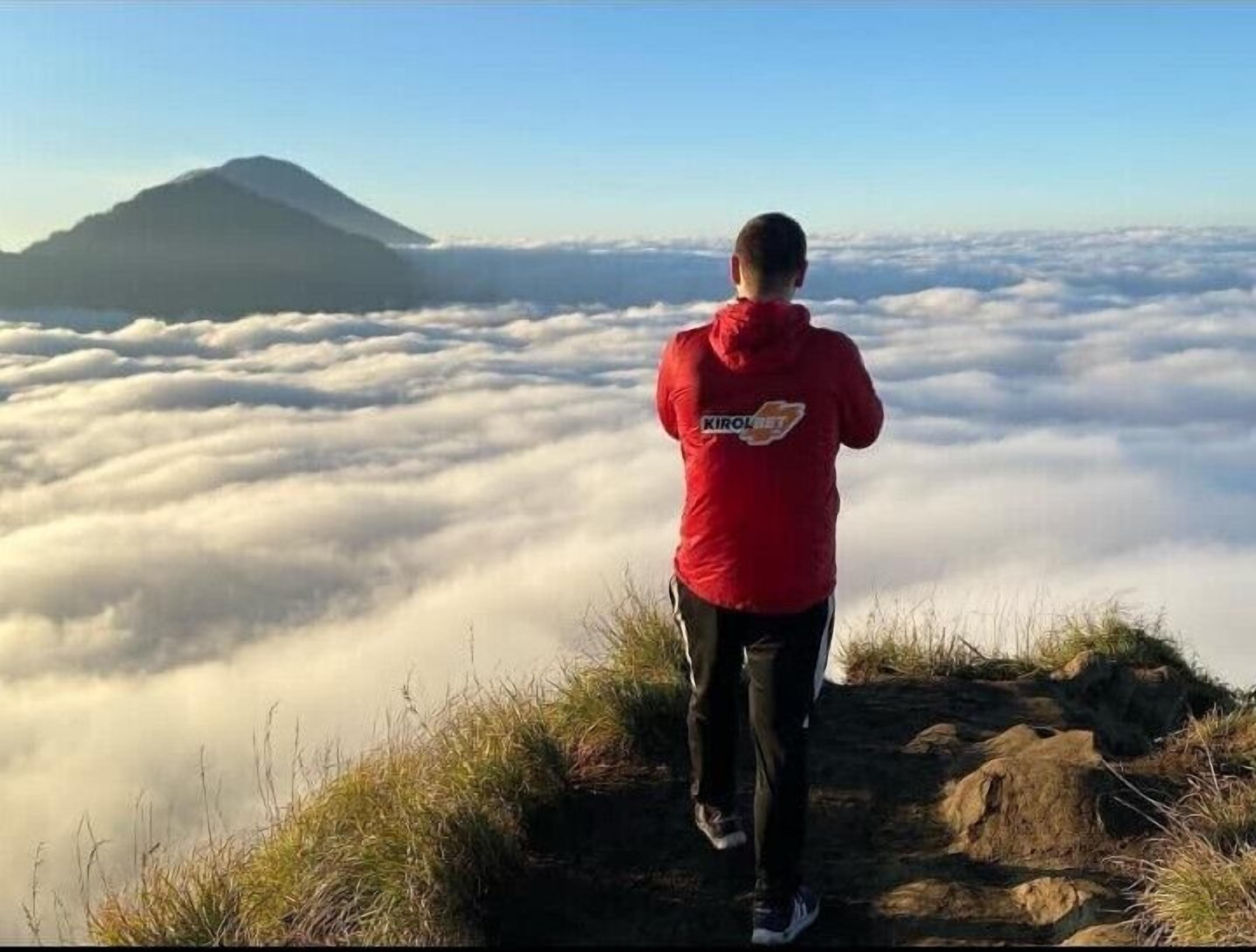 A person in a red jacket standing on a rocky outcrop overlooking a sea of clouds with a mountain in the distance.