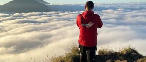 A person in a red jacket standing on a rocky outcrop overlooking a sea of clouds with a mountain in the distance.