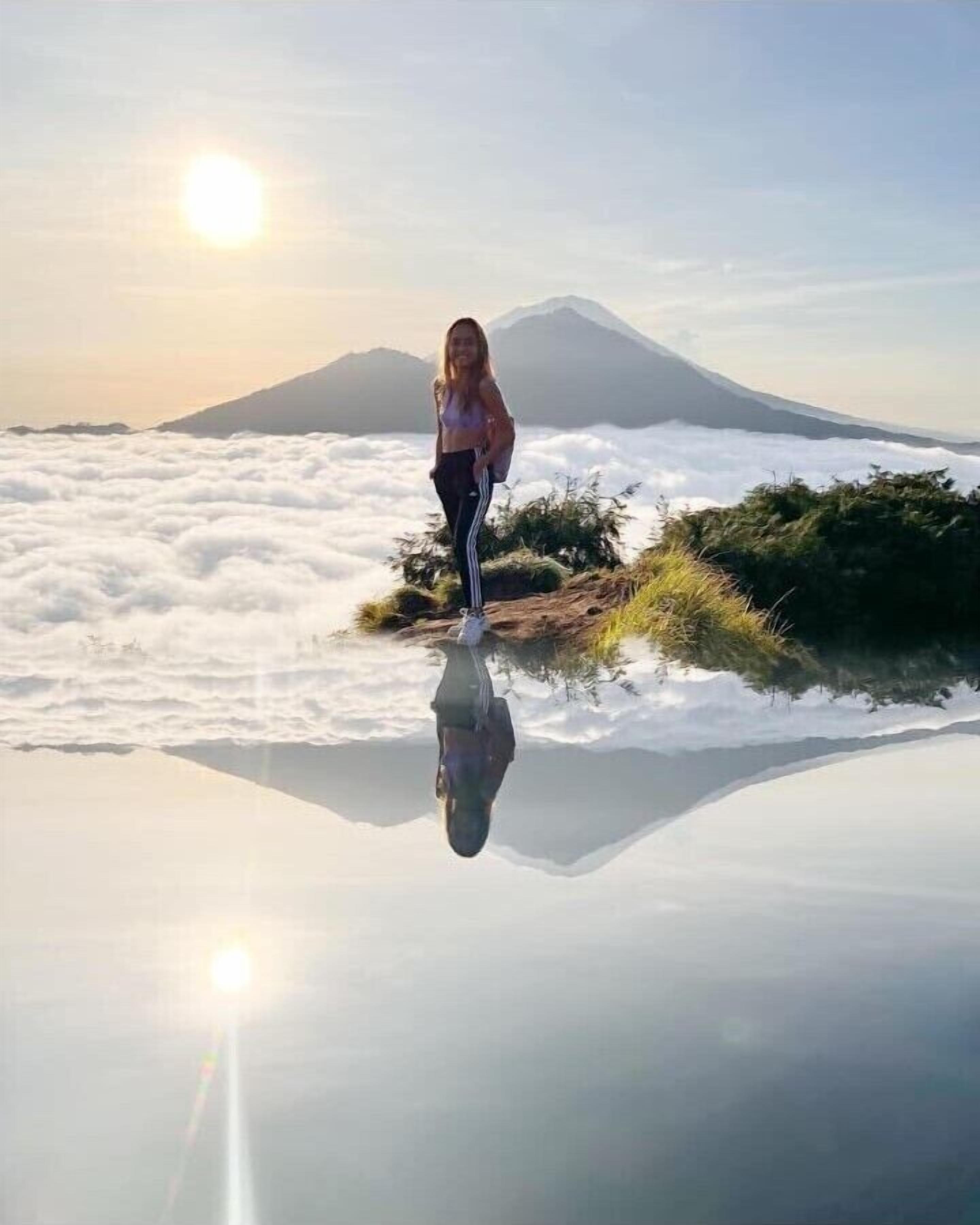 A person standing on a reflective surface with a mountain and clouds in the background.