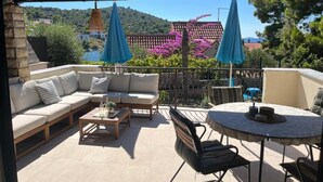 An outdoor patio with a sofa, chairs, and a table. There are two umbrellas and a view of a hillside with buildings and trees. - Holiday house Mia (Rogoznica)