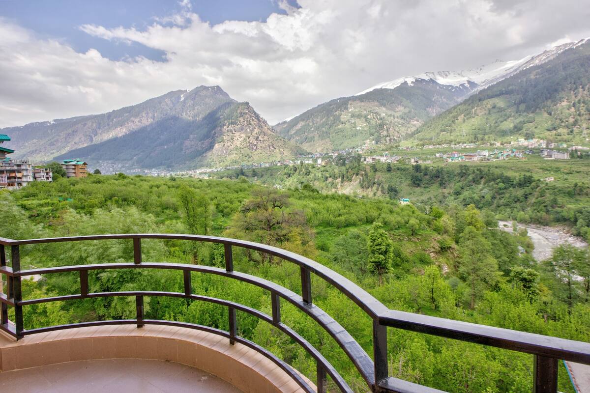 A scenic view from a balcony with a railing, overlooking a valley with mountains, a river, and a small town.