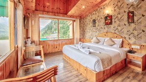 A wooden cabin room with a large bed, a wooden chair, a table with a glass top, and a view of the mountains through a large window. - Hotel Nirosha Manali (Manali)