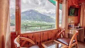 A wooden cabin with a large window offering a view of a mountainous landscape. Inside, there is a wooden table and two chairs. - Hotel Nirosha Manali (Manali)
