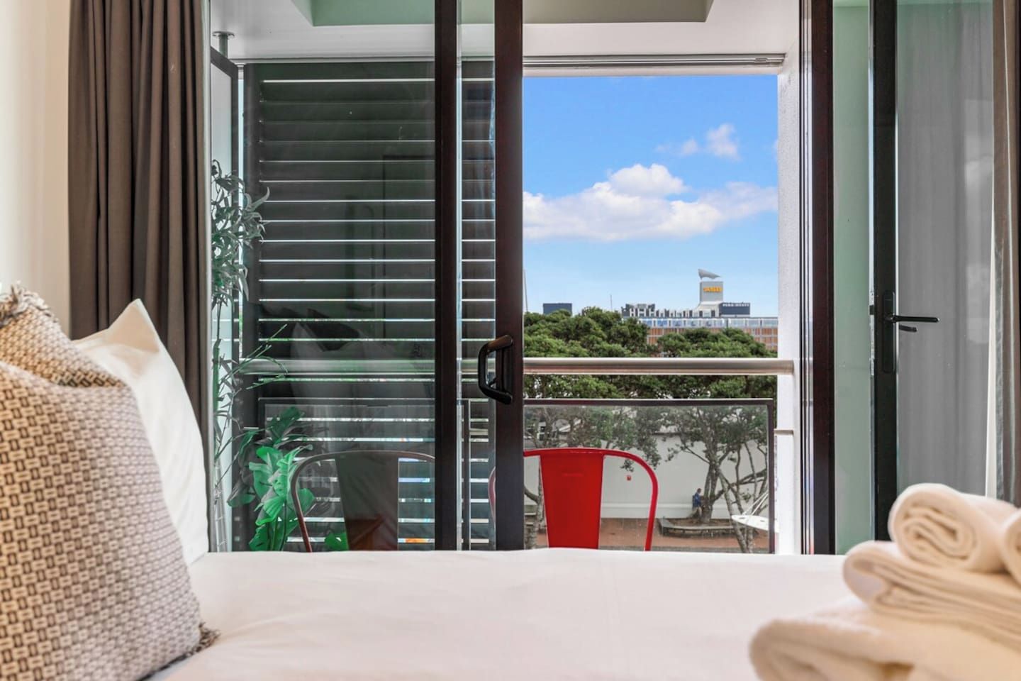 A hotel room with a bed, a view of the city through the window, and a balcony with a chair and a plant.