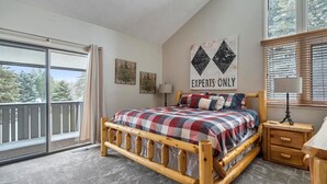 Un dormitorio con una cama de madera, una mesita de noche, un ventanal con persianas y un balcón con vistas a árboles.