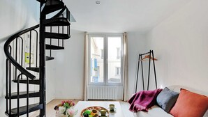 Ein modernes Wohnzimmer mit einer Wendeltreppe, einem Esstisch mit Obstschale und Tassen sowie einem Sofa mit Kissen.