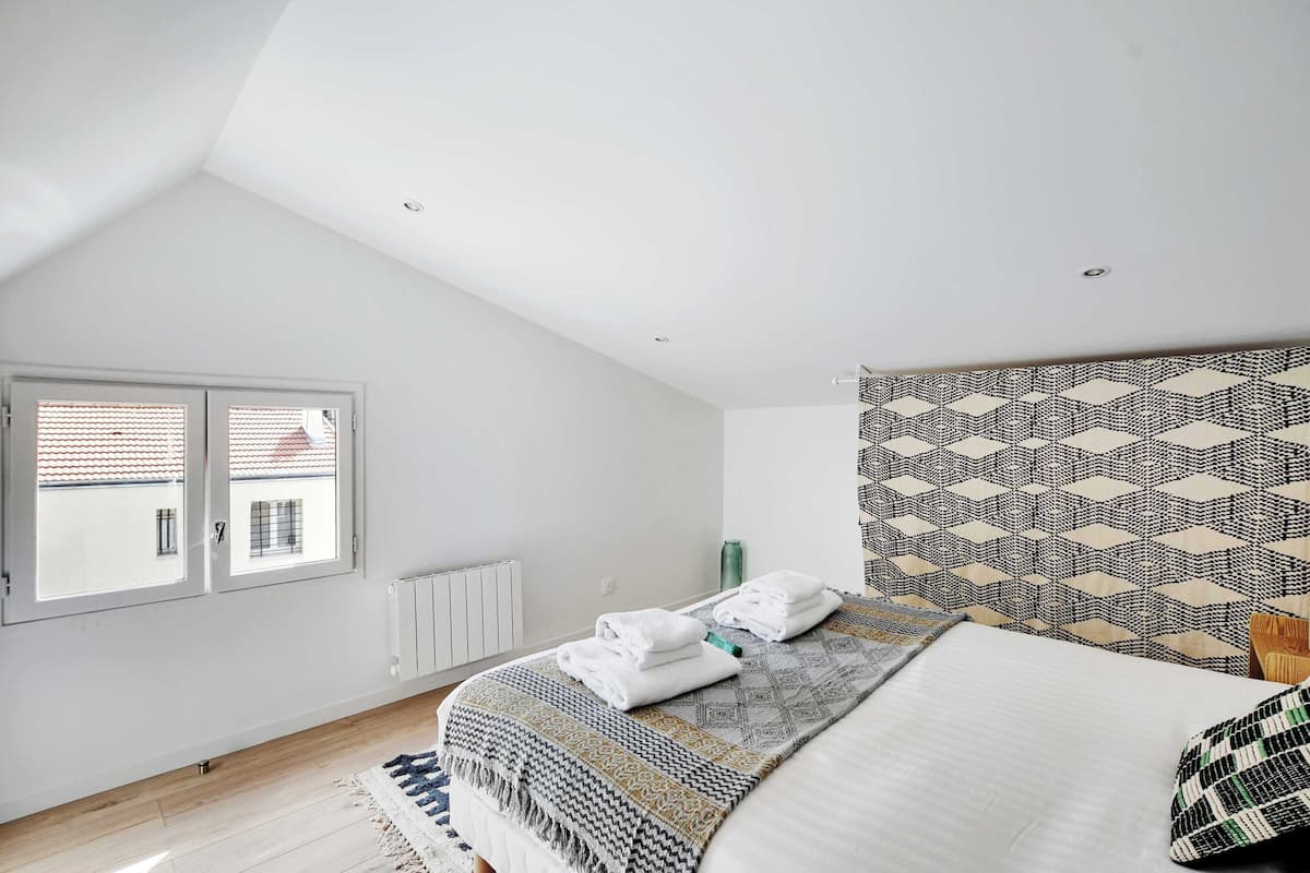 Ein Schlafzimmer mit schrägen Wänden, einem Bett mit gemustertem Kopfteil, einem Fenster mit Jalousien und einer Heizung.