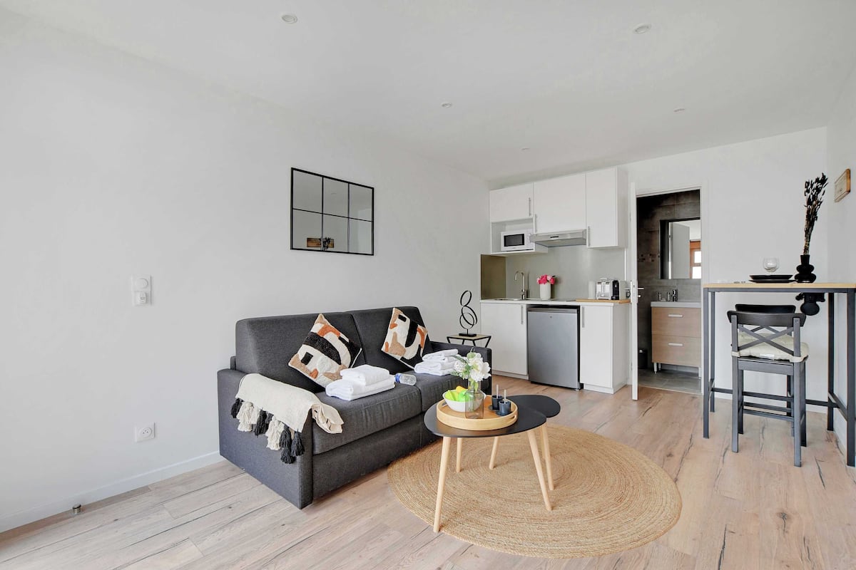 Ein modernes Wohnzimmer mit einem grauen Sofa, einem kleinen runden Tisch mit Tablett und einer Küchenzeile mit weißen Schränken und einer Mikrowelle.