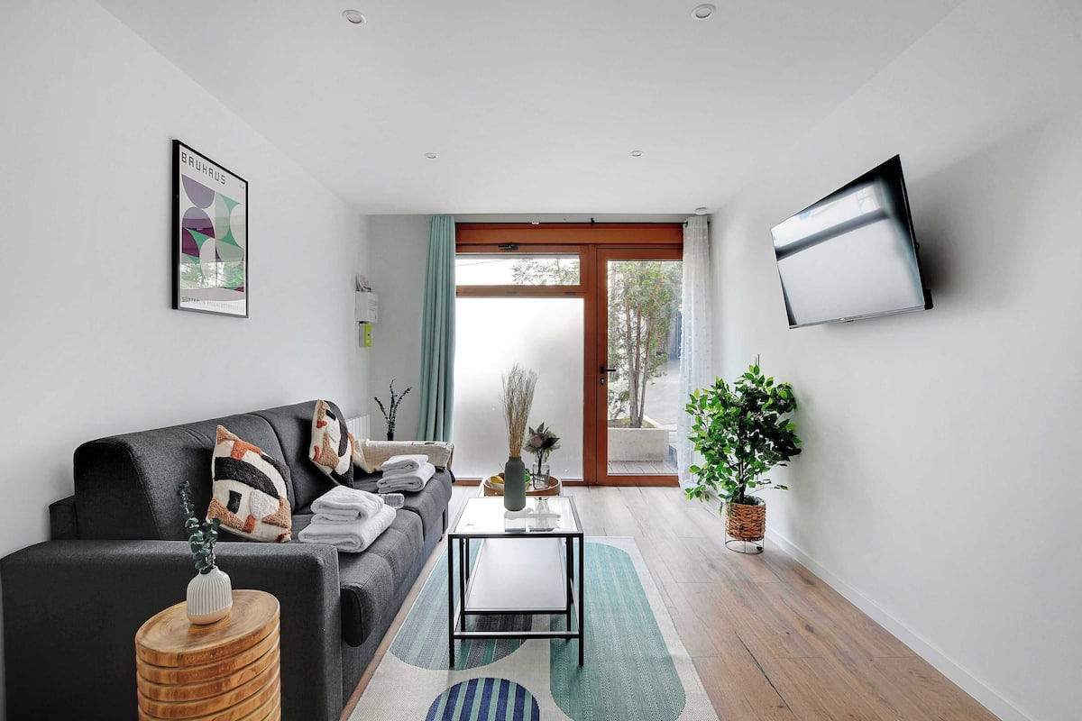Ein modernes Wohnzimmer mit einer Couch, einem Couchtisch, einem an der Wand montierten Fernseher und einem großen Fenster mit Blick auf Bäume.
