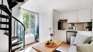 Eine moderne Küche mit weißen Schränken, einer Mikrowelle und einem kleinen Kühlschrank. Ein Esstisch mit Obstschale und Blumenvase steht daneben. Eine Wendeltreppe ist links zu sehen.