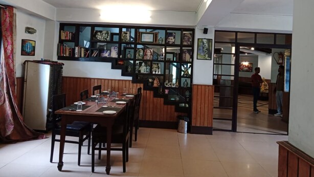 A dining area with a long wooden table set for four, a wall-mounted bookshelf with various items, and a doorway leading to another room. - Hotel Dew Pond  (Gangtok)
