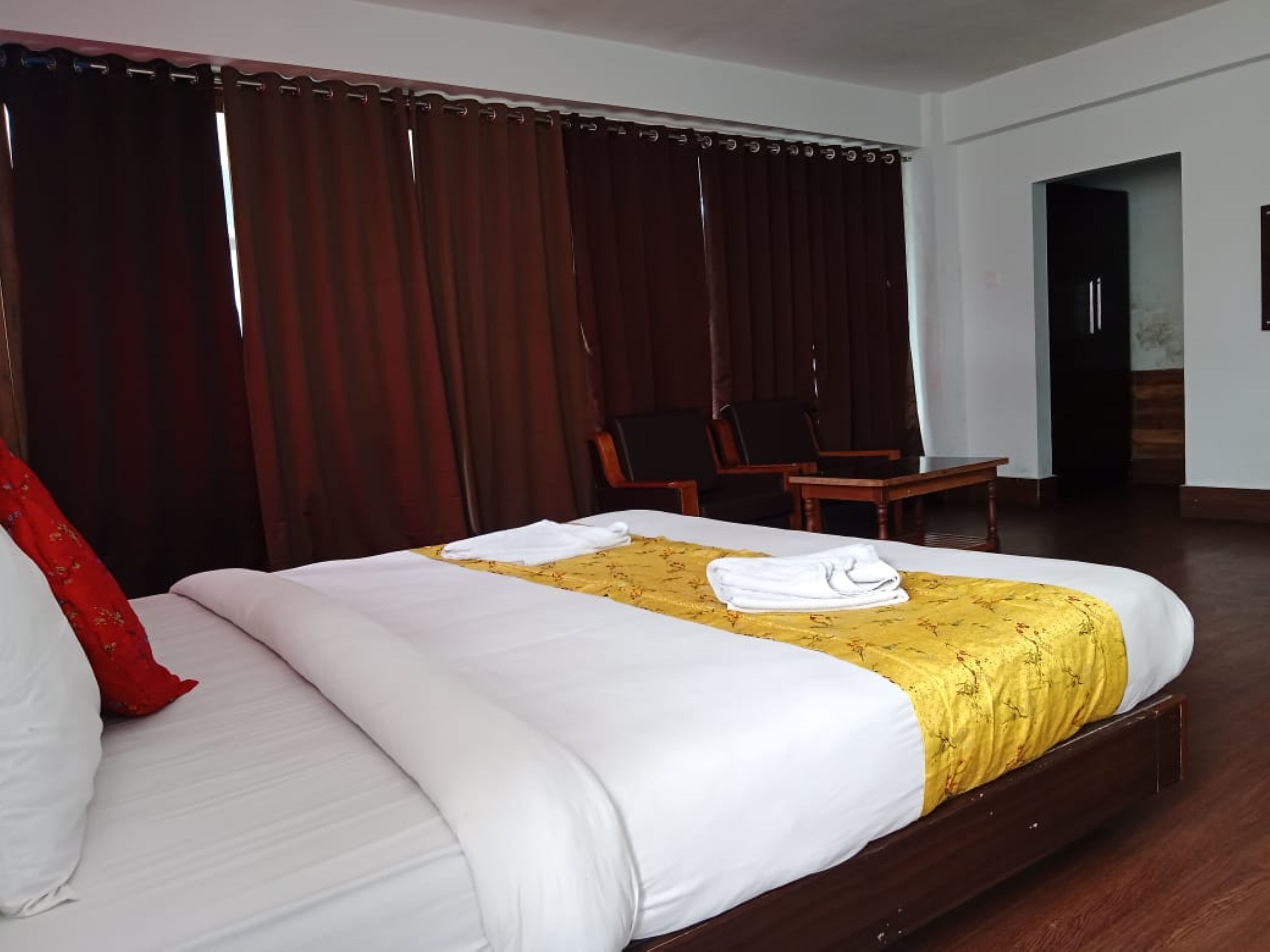 A hotel room with a large bed, a wooden headboard, a yellow bedspread, and a red throw pillow. There is a chair and a small table in the corner.