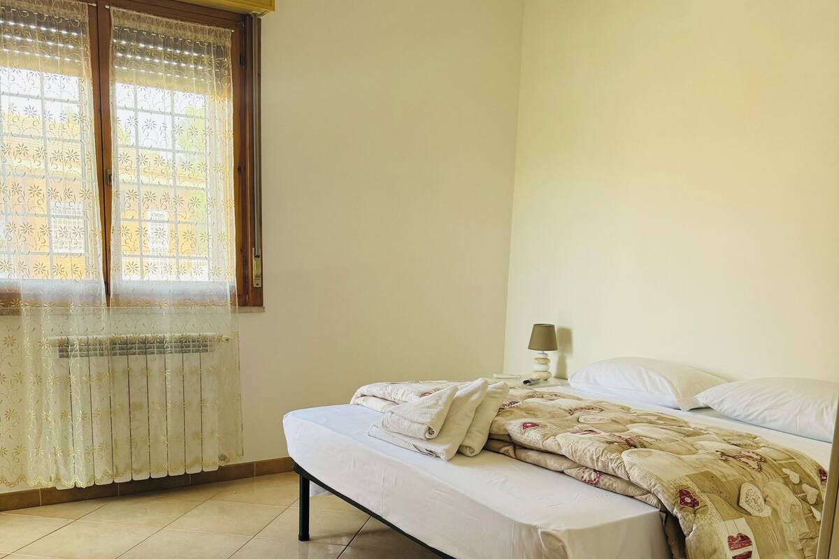 A bedroom with a bed, a nightstand, a lamp, and a window with patterned curtains.