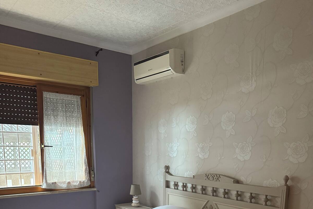 A neatly made bed with a floral bedspread, a bedside lamp, and a wall-mounted air conditioner.