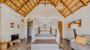 A bedroom with a bed under a thatched roof, a sofa, and a television. - Zaria Garden Bungalow (Pwani Mchangani)