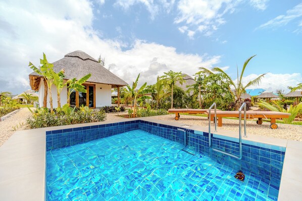 A swimming pool with a tiled edge, surrounded by palm trees and a thatched-roof building. - Zaria Garden Bungalow (Pwani Mchangani)
