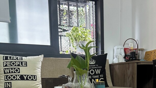 Une table en bois sur laquelle se trouvent un vase rempli de plantes vertes, une petite théière et une tasse. Un coussin orné d’une citation inspirante et une fenêtre donnant sur un paysage verdoyant complètent la scène.