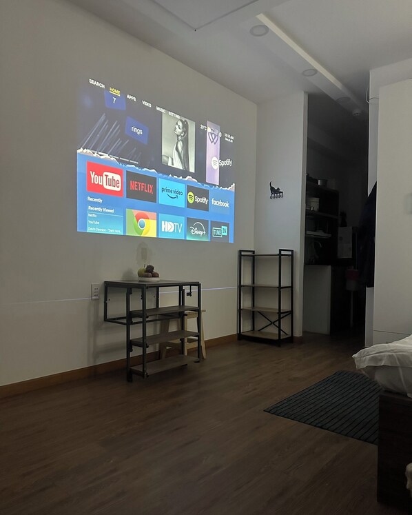 A modern room with a wall-mounted TV displaying various streaming services. There is a small table with a lamp and a shelf unit. The room has wooden flooring and a white door. - Tom's Homestay (Ho Chi Minh City)
