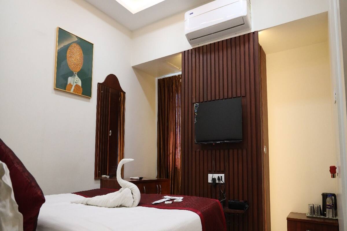 A hotel room with a bed, a flat-screen TV mounted on the wall, an air conditioning unit, a wooden wardrobe, and a painting on the wall.