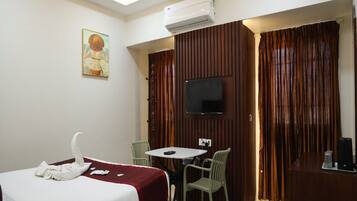 A hotel room with a bed, a dining table with chairs, a television, and an air conditioning unit.