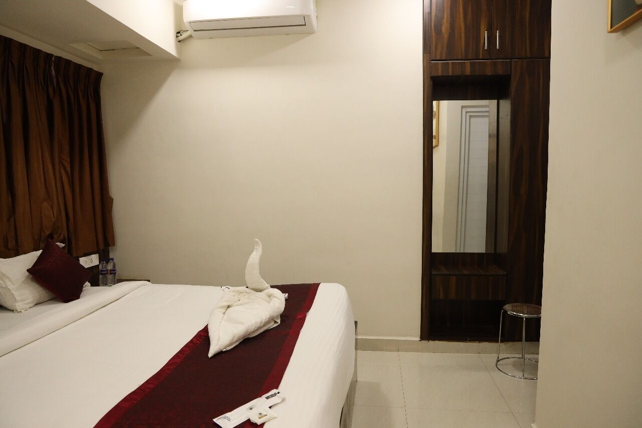 A hotel room with a bed, a red blanket, a white towel, a wooden wardrobe, and a small stool.