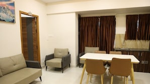 A living room with a sofa, two armchairs, a dining table with four chairs, a kitchen area with a sink, and a large window with curtains. - Manopriya Residency by Azalea (Madurai)