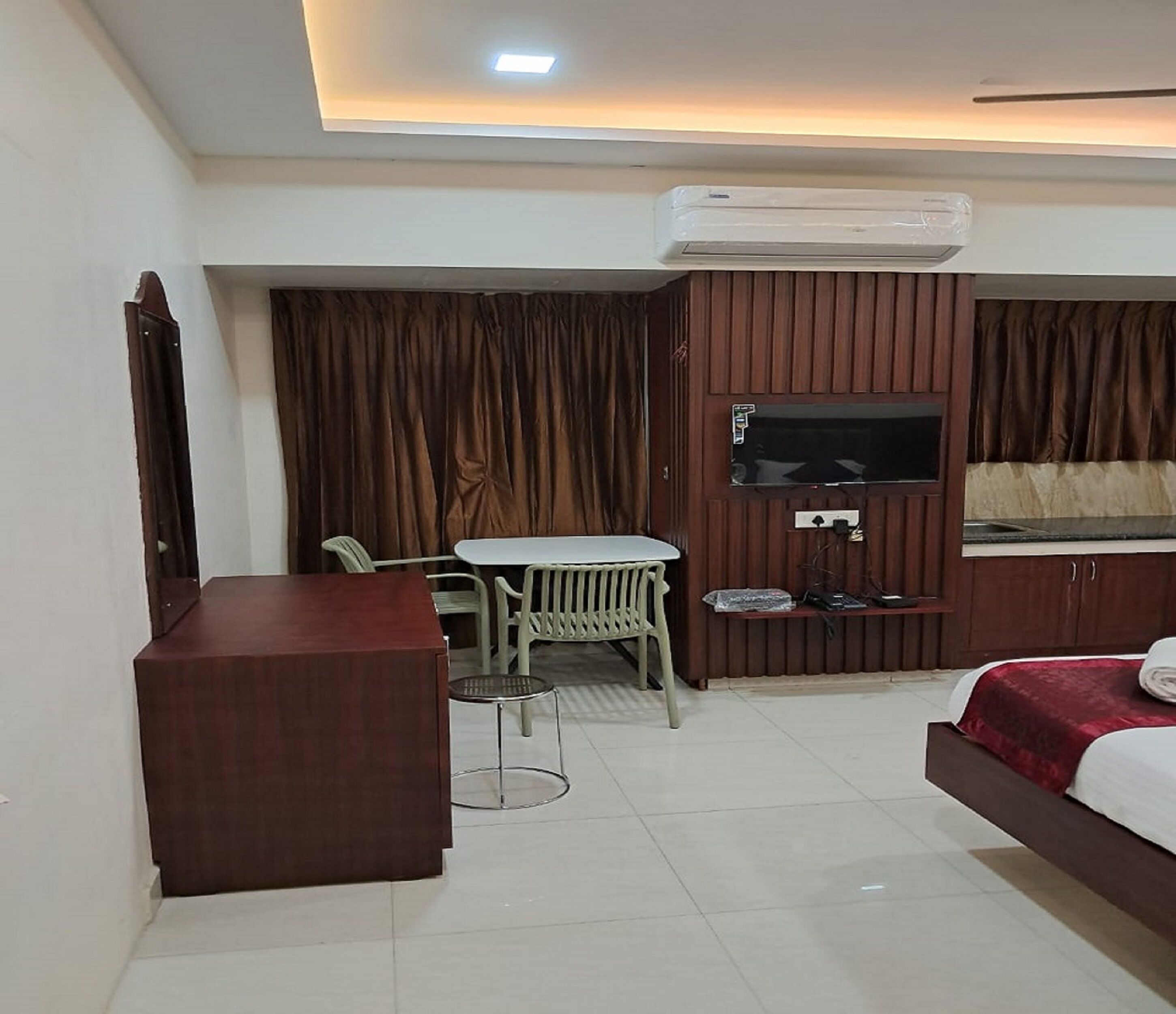 A hotel room with a bed, a desk, a chair, a TV mounted on the wall, and an air conditioning unit.