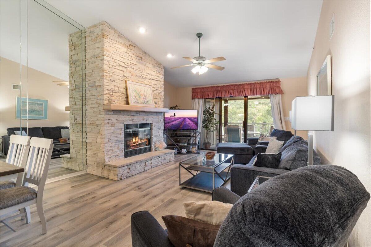 A living room with a stone fireplace, a ceiling fan, and a large window with a view of greenery.
