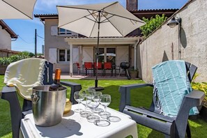 En terrasse med havemøbler, et bord med en flaske og glas, og et hus i baggrunden.