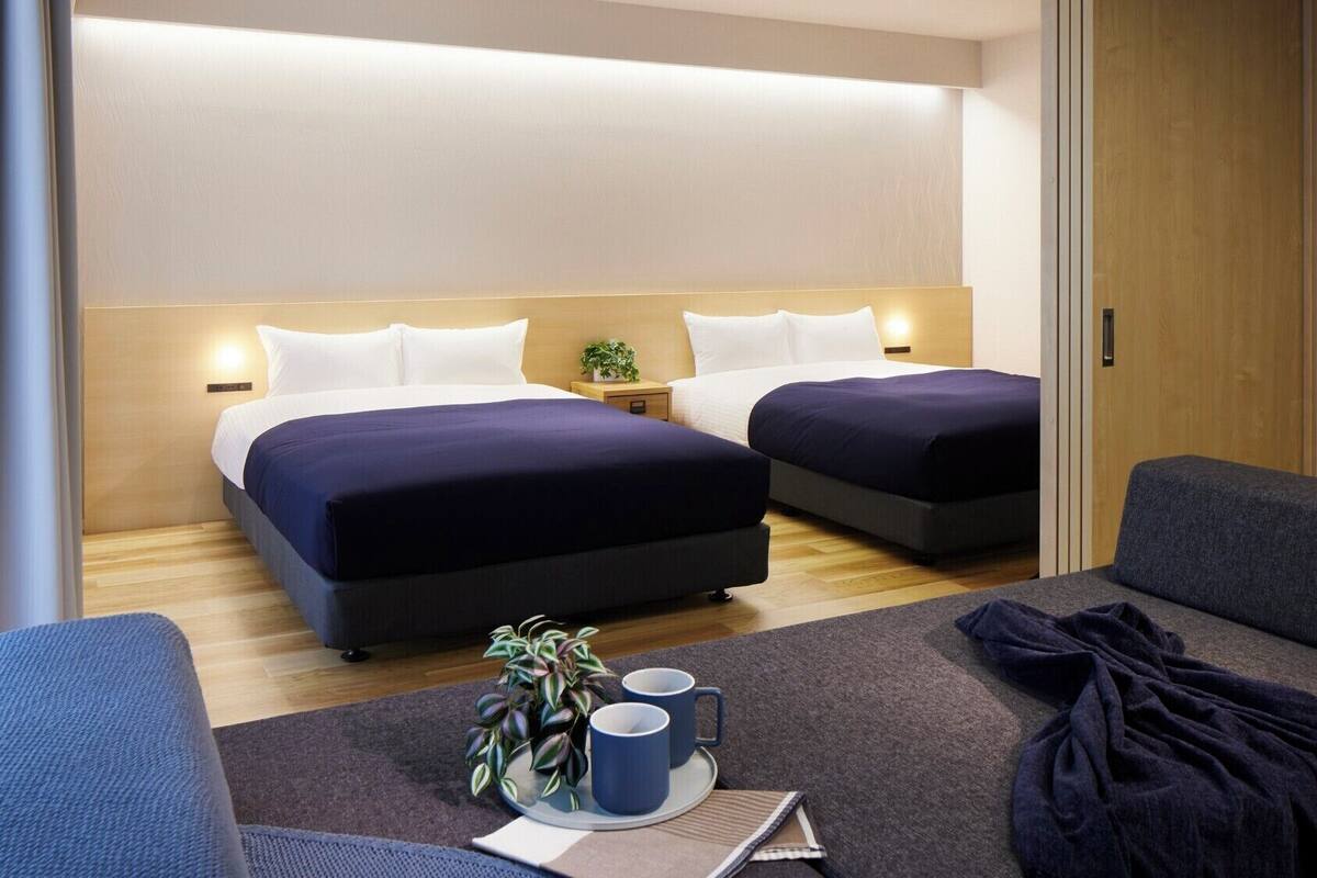 Une chambre d’hôtel avec deux lits, un canapé, une petite table sur laquelle se trouve un plateau avec divers objets, et une plante en pot.