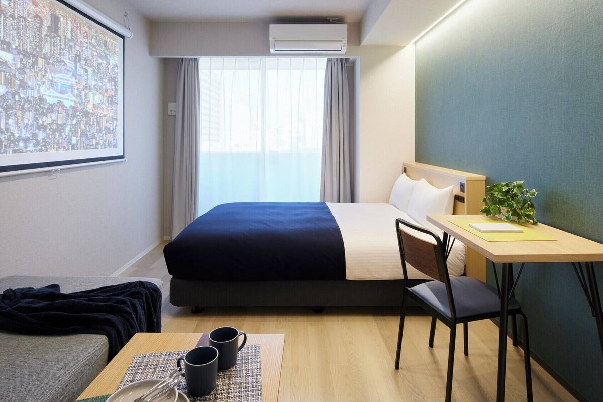 Une chambre d’hôtel équipée d’un lit, d’un bureau, d’une chaise, d’un canapé, d’une table basse et d’un écran de projection diffusant une vue sur la ville.
