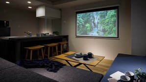 A cozy living room with a kitchen area, a projector screen displaying a forest scene, and a comfortable seating arrangement.