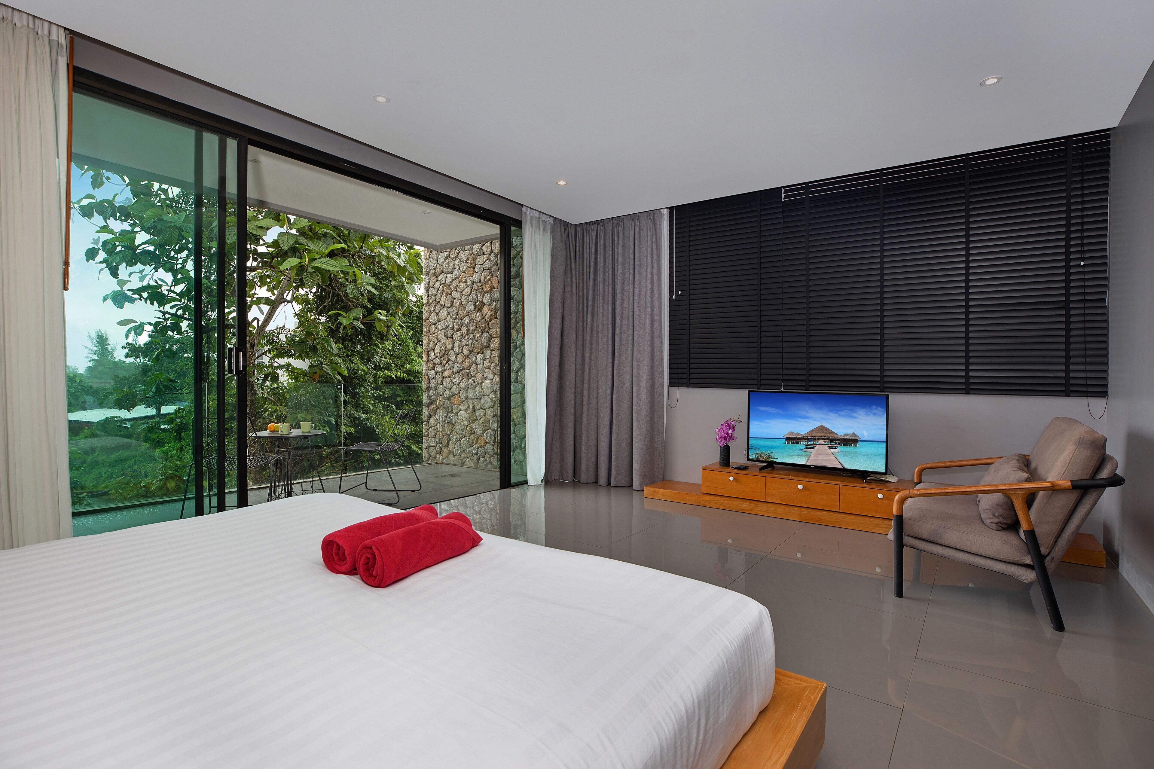 A modern hotel room with a large bed, a TV, a seating area, and a view of greenery.