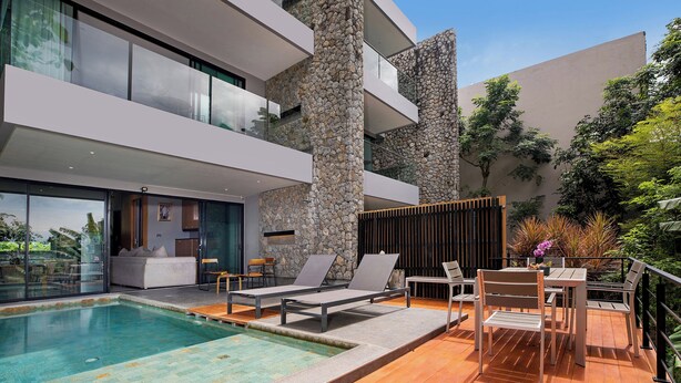 A modern outdoor pool area with lounge chairs and a table, surrounded by a wooden deck and stone walls. - Designer 4BR Pool Villa Gentle Breeze (Kamala)