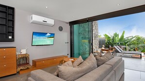 A modern living room with a large sofa, a television, and a view of lush greenery outside. - Designer 4BR Pool Villa Gentle Breeze (Kamala)