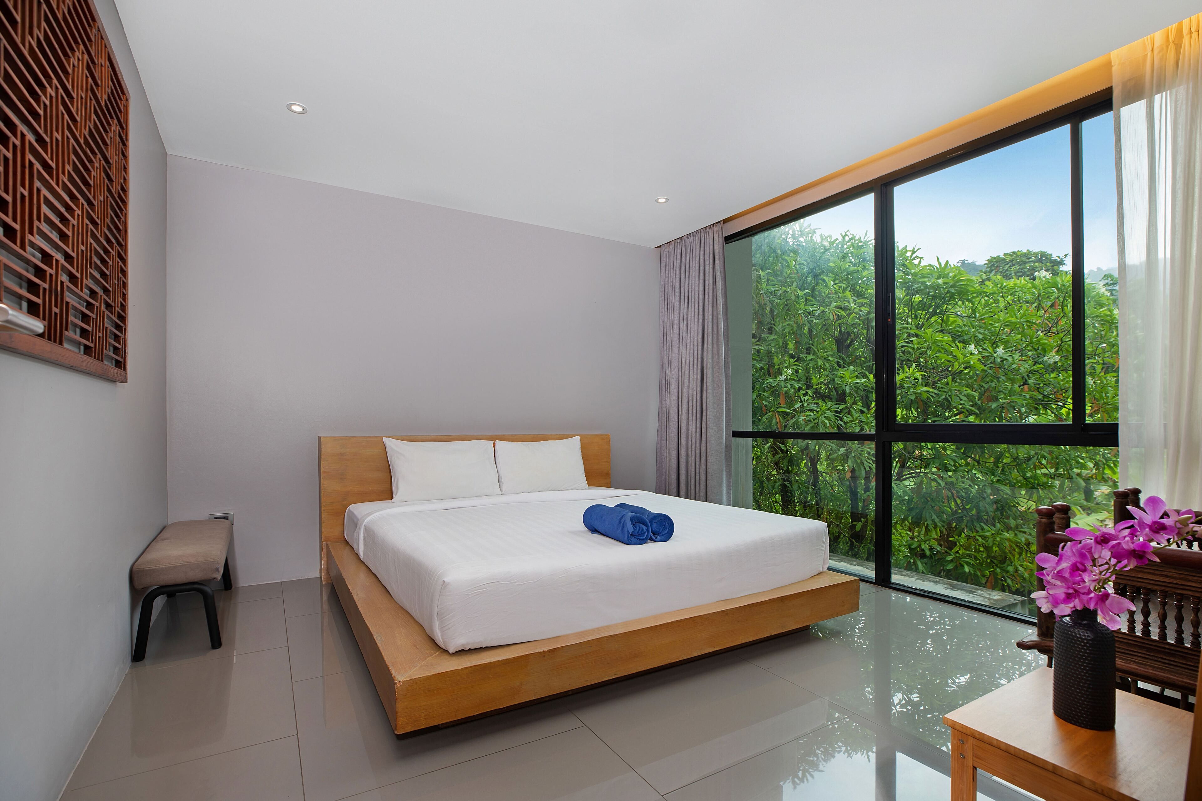 A modern hotel room with a large bed, a small bench, a table with a vase, and a view of greenery through large windows.